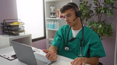 Kulaklığı ve steteskopu olan yeşil önlüklü genç adam klinikteki bir masada çalışıyor. Organize bir hastane odasında tele-sağlık danışmanlığı için dizüstü bilgisayar kullanıyor..