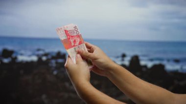 Hong Kong banknotlarını sahil şeridinde tutan bir adam şehir arkaplanı ve deniz dalgaları ile açık havada finansal yatırım sergiliyor ve iş odaklı bir atmosfer yaratıyor..