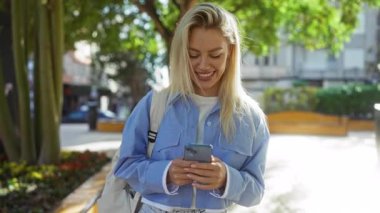 Güneşli şehir parkında canlı bir günde ağaçlar ve çiçeklerle akıllı telefonlara bakıp gülümseyen bir kadın.