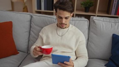 Genç adam oturma odasında bir kanepede oturuyor, dijital tablet kullanırken kırmızı bardaktan kahve içiyor, rahat, şık ve modern bir atmosfer yayıyor..