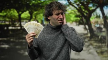 Parkta Norveçli kronu tutan İspanyol bir adam, doğal ortamda kulak ağrısını dile getiriyor, maddi rahatsızlığı ya da stresi temsil ediyor..