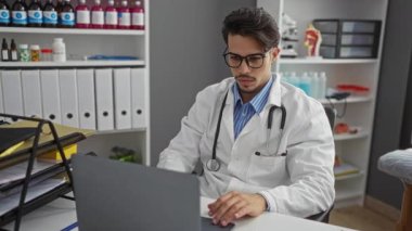 Klinik ortamındaki genç adam, tıbbi malzemeler ve organize raflarla çevrili laboratuvar önlüğü ve steteskop giyerken dizüstü bilgisayarına odaklanıyor..