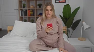 Sarı saçlı hamile bir kadın rahat bir yatak odasında bir yatakta otururken üzgün görünüyor, akıllı telefonu içeride tutuyor, rahat bir ev ortamında hayal kırıklığını ifade ediyor..