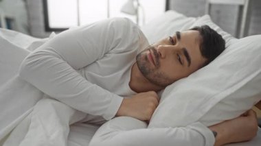 Yatakta gülümseyen genç adam, yatak odası, doğal ışık ve rahat atmosfer, beyaz gömlek giyiyor..