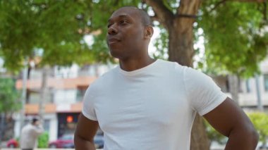 Yeşil bir şehir parkında gülümseyen bir adam, elleri kalçasında, beyaz bir tişört giymiş, rahat bir sokak ortamının özünü yakalıyor..