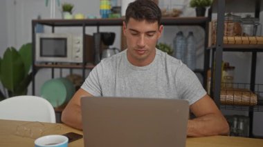 Laptopta çalışan genç İspanyol bir adam içi bitki ve mutfak malzemeleriyle dolu rahat bir ofis ortamında düşünceli ve elimizdeki işe odaklanmış görünüyor..