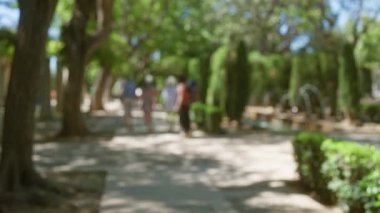 Mallorca 'da yemyeşil ağaçları ve güneşli bir atmosferi olan açık hava parkında yürüyen odaksız insanlar.