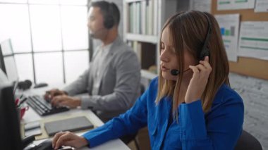 Modern ofis ortamında bilgisayar başında çalışan kadın ve erkek, kulaklık takıyor, görevlere odaklanıyor, iş ortamında takım çalışmasını ve iletişimi temsil ediyor..