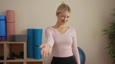 Arka planda yoga minderleri ve ekipmanlarla bir spor salonunda güvenle poz veren kadın, dostça bir gülümseme ve rahat bir duruş sergiliyor..