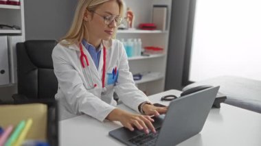 Klinikte dizüstü bilgisayarda steteskop giymiş, gülümseyen organize klinik çalışma alanı rafları dizüstü bilgisayarı olan bir kadın.