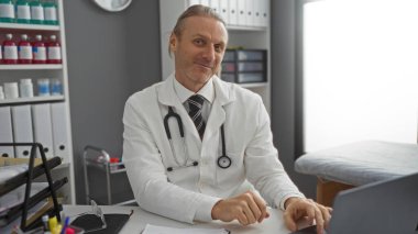 Steteskopla hastane ofisinde oturan olgun sarışın beyaz erkek doktor, düzenli bir klinik ortamında güvenle gülümsüyor, profesyonelliği canlandırıyor.