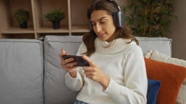 Kulaklık ve süveter takan bir kadın koltukta rahat bir şekilde akıllı telefon kullanıyor. Çiçekler ve renkli yastıklarla dekore edilmiş oturma odasında..