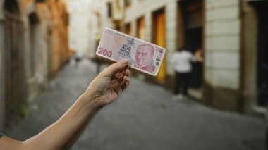 Bir şehir caddesinde Türk lirası banknotunu tutan bir adam, para ve arka plan bulanıklığı odaklı şehir manzarası çekiyor..