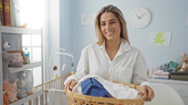 Çamaşır odasında elinde sepetle gülümseyen bir kadın. Etrafı yumuşak oyuncaklar ve ev ortamında çocuk odası dekoruyla çevrili..