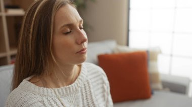 Rahat bir oturma odasında, gözleri kapalı, beyaz bir süveter giyen, huzurlu bir ortam sergileyen, düşüncelere ve sakinliğe önem veren bir kadın..