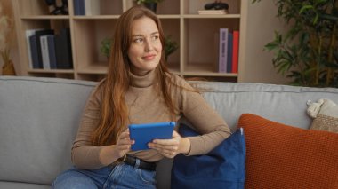 Tabletli bir kadın, rahat, modern bir oturma odasında, rahat bir atmosfer sergiliyor. Şık bir dekor ve doğal ışık, teknoloji ve yaşam tarzı teması için ideal..