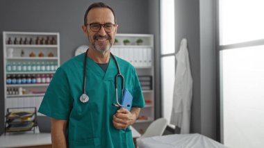 Önlüklü İspanyol bir adam hastane odasında güvenle gülümsüyor. Tıp alanında profesyonelliği ve bakımı sembolize ediyor..