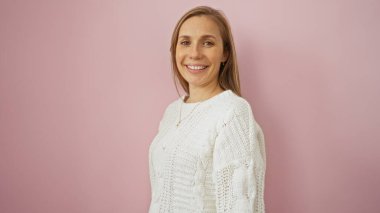 İzole edilmiş pembe bir arka plana gülümseyen, rahat beyaz bir kazak giyen, stüdyo ortamında neşeli bir ifade yakalayan bir kadın..