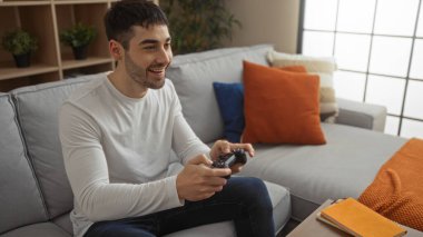 Genç adam rahat bir oturma odasında video oyunları oynuyor, gülümsüyor ve odaklanıyor, minderlerle ve boş eşyalarla çevrili, rahat ve neşeli bir atmosfer yaratıyor..