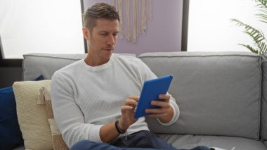 Evdeki kanepede rahat rahat oturan bir adam, rahat bir oturma odasında rahat bir şekilde giyinmiş, parlak dekorlu ve yumuşak yastıklı, boş zamanların keyfini çıkarıyor..