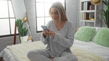 Akıllı telefon kullanan bir kadın, rahat bir yatak odasında, yumuşak yastıklarla dekore edilmiş, modern dekorasyon ve geniş pencerelerden yansıyan doğal ışık ile çevrili, huzurlu bir yaşam tarzı sergiliyor..