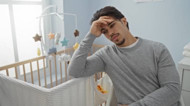 Genç İspanyol bir adam yatak odasında yorgun görünüyor. Bebek beşiğinin yanında oturuyor. Günlük giysiler giyiyor ve düşünceli bir ifadeyle başını uzatıyor..