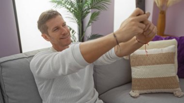 Modern oturma odasında selfie çeken genç bir adam şık dekorlu rahat koltukta oturuyor, rahat bir yaşam tarzı ve çağdaş bir ev ortamı sergiliyor..