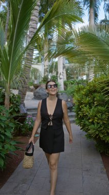 Lanzarote 'de, kanarya adalarında gezen, gülümseyen, güneş gözlüğü takan ve yemyeşil palmiye ağaçları ve tropikal bitkiler arasında hasır bir çanta tutan genç bir kadın.