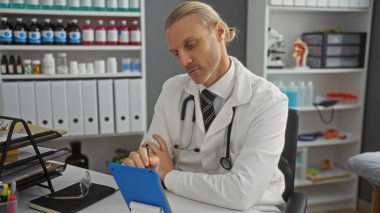 Klinikteki orta yaşlı sarışın adam beyaz önlük ve steteskop giyiyor. Masasında raflar ve gözlüklerle iyi organize edilmiş bir muayenehanede tablete konsantre oluyor..