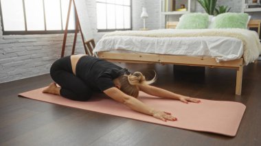Konforlu bir yatak odasında, ahşap döşemeli ve modern dekorlu yoga yapan bir kadın dinlendirici egzersiz ve farkındalık egzersizi için sakin bir iç mekan ayarını sergiliyor..