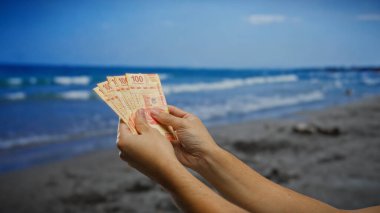 Sahil kenarında Meksika pezosu tutan bir adam, deniz kenarı ortamında finansal yatırımın özünü yakalıyor, seyahat ve para alışverişini vurguluyor..