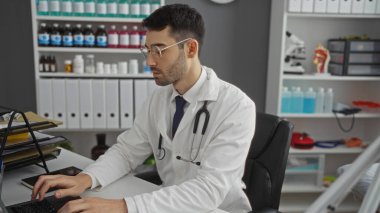Hastane kliniğinde çalışan, steteskoplu laboratuvar önlüğü giyen genç bir adam, organize bir iç mekanda tıbbi malzemelerle çevrili bilgisayar görevlerine odaklanmış.