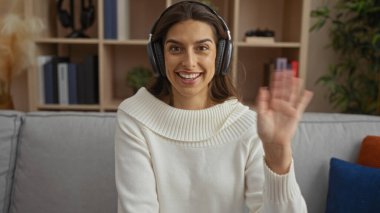 Yüzünde kulaklıkla gülümseyen bir kadın oturma odasında modern dekorasyon ve teknoloji nişanını sergiliyor.