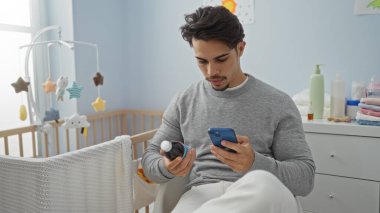 Yatak odasındaki genç adam telefon ve ilaç şişesi tutuyor, rahat bir ev ortamında, yanında beşikle, kapalı bir ortamda özenli bakım gösteriyor..