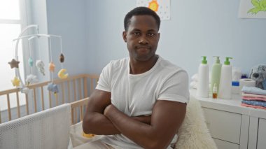 Çapraz kollu genç adam beşikteki rahat bir yatak odasında oturuyor, yumuşak mobilyalar ve yumuşak renklerle çevrili, huzurlu ve sıcak bir ev ortamı yaratıyor..