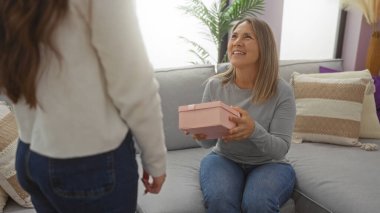 Anne, oturma odasındaki kanepede kızından pembe bir hediye alıyor ve evde aile sevgisini ifade ediyor.