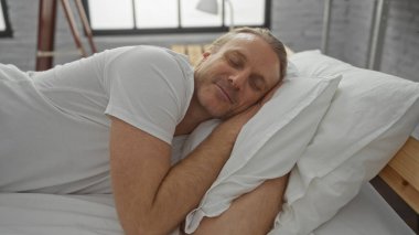 Olgun sarışın bir adam, rahat bir yatak odasındaki bir yatakta huzur içinde uyur. Rahat ve rahat bir iç dekorasyon sergiler..