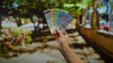 Hand of man holding malaysian ringgit banknotes in an outdoor urban park setting, emphasizing business and finance in a city environment. clipart