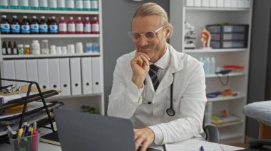 Modern bir klinikte çalışan doktor, orta yaşlı, beyaz, sarı saçlı, gözlüklü, etrafı tıbbi malzeme ve ekipmanlarla çevrili, dizüstü bilgisayar kullanan bir adam..