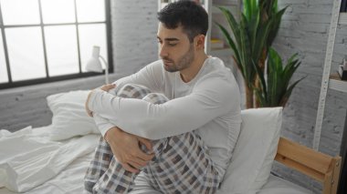 Genç İspanyol adam yatak odasında, yumuşak ışıkla düşünceli görünüyor, rahat iç ve yeşil bitkilerle çevrili, rahat pijamalar giyiyor..