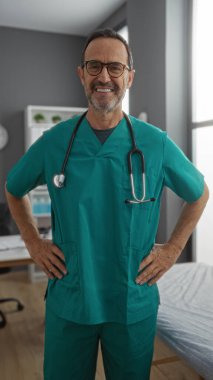 Yeşil önlüklü, olgun İspanyol bir adam klinik odasında güvenle gülümsüyor. İçerideki hastane ortamında sağlık hizmetleri profesyonelliğini ve hasta bakımını temsil ediyor.