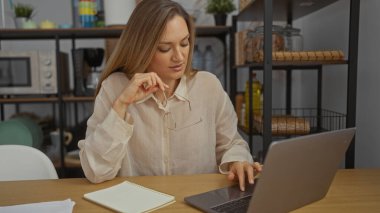 Ofis masasında dizüstü bilgisayar, defter ve modern iş ortamında düşünceli bir ifadeyle çalışan bir kadın profesyonel iç mekandaki görevine odaklanıyor..