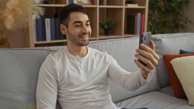 Genç İspanyol bir adam rahat bir oturma odasında kanepede oturmuş selfie çekiyor ve evlerinin sıcaklığını ve rahatlığını gösteriyor..