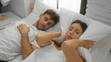 Kadın yastıkla kulaklarını kapatırken, erkek modern yatak odasında huzur içinde uyuyor ve samimi ev ortamını gözler önüne seriyor..