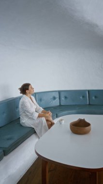 Beyaz elbiseli genç bir kadın Mirador del Rio, Lanzarote, Kanarya Adaları 'nda oturuyor. Mavi yastıklı koltukları ve kilden bir kasesi olan beyaz bir masası var.