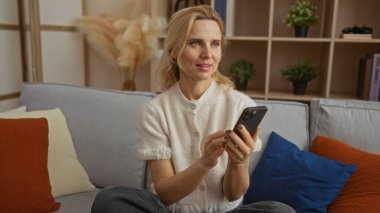 Elinde telefon tutan bir kadın şık dekorlu rahat bir oturma odasında rahat bir koltukta oturuyor. Parlak ve davetkar bir atmosfer sergiliyor..