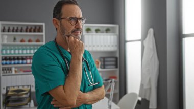 Tıbbi ekipman ve malzemelerle çevrili bir hastane odasında steteskopla önlük giyen yetişkin bir İspanyol, klinik bir ortam sergiliyor..