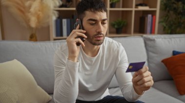 Oturma odasında kredi kartıyla konuşan genç adam düşünceli ve odaklanmış görünüyor..