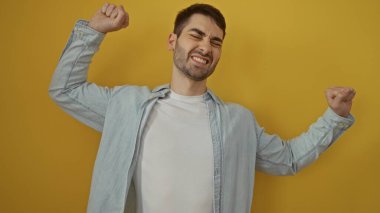 Genç adam neşeli bir şekilde sarı bir arka plana yaslanıyor, kendine güven ve enerji yayıyor, dinamik bir portre yaratıyor çekiciliği ve pozitif titreşimleri vurguluyor..