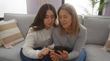 Anne ve kızı rahat bir oturma odasında bir kanepede oturuyor, aile sevgisi ve bağlantısını paylaşıyor, akıllı telefon ekranına odaklanıyor, birbirlerinin arkadaşlığından keyif alıyorlar..